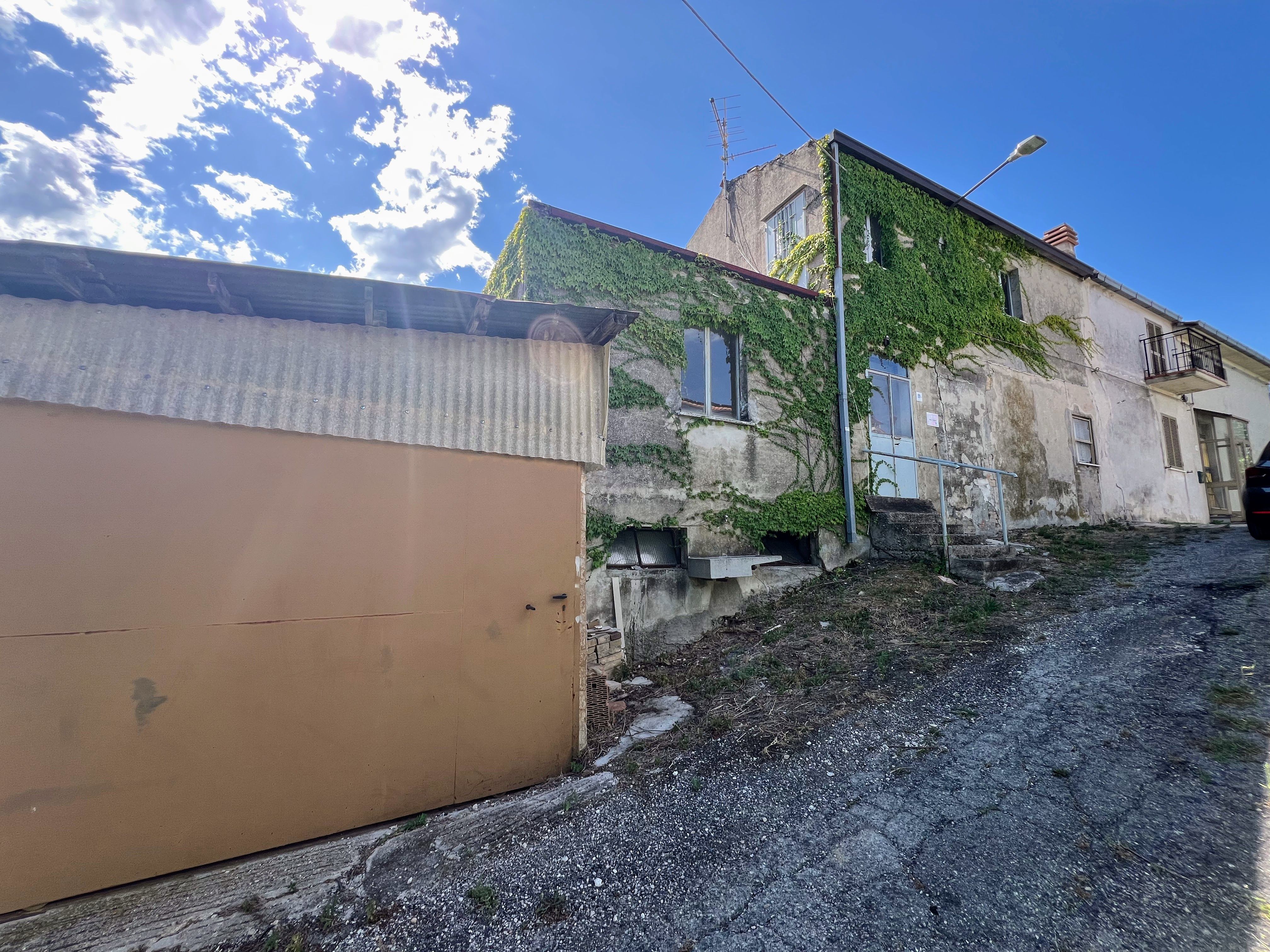 Ivy-covered roadside house in Abruzzo, viewed uphill from a gravel lane, with a corrugated garage structure, stone walls, a central doorway and a streetlight.