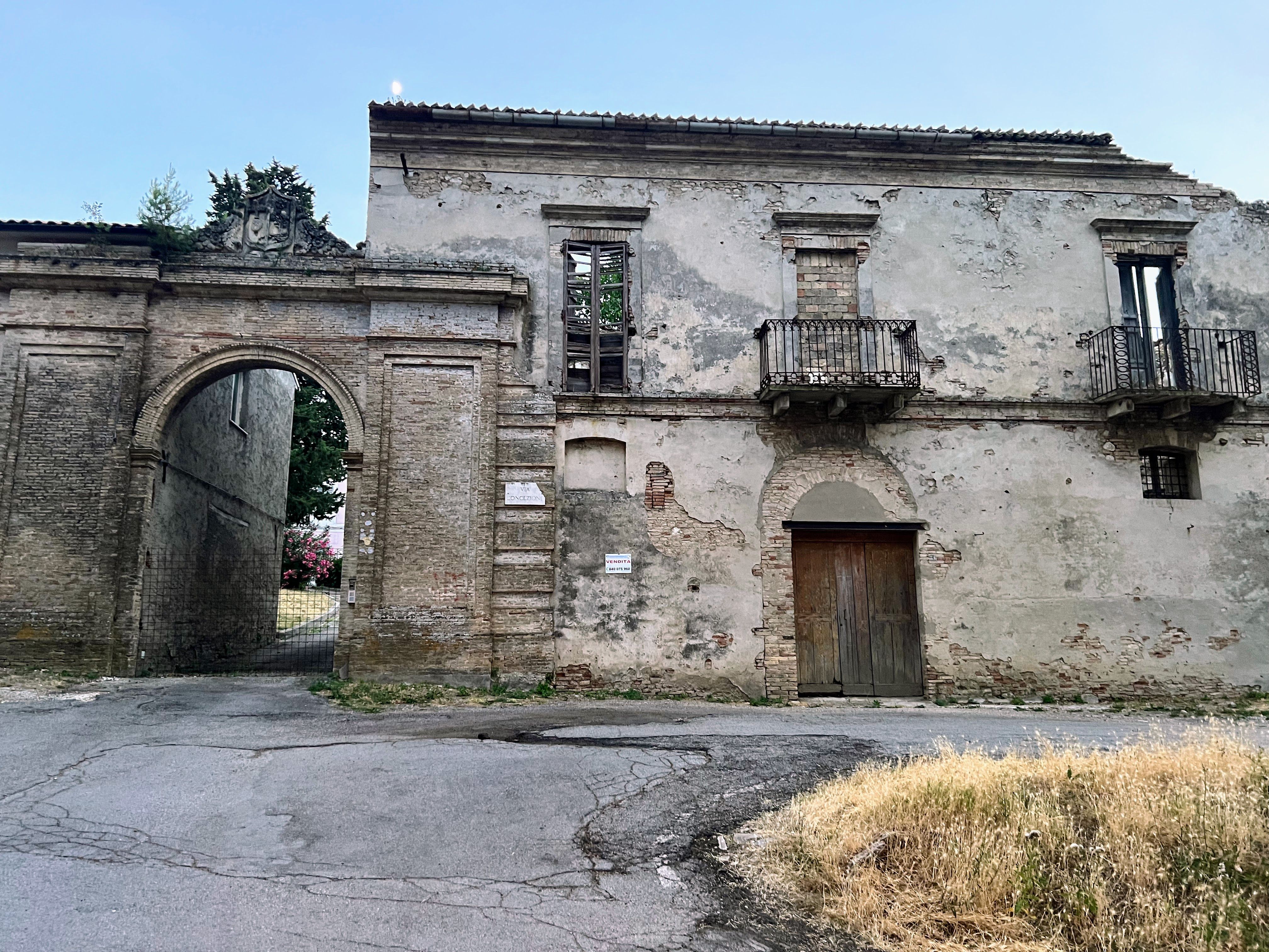Original photograph of a weathered large historic building in Abruzzo with an arched gateway, boarded openings and cracked road in front.