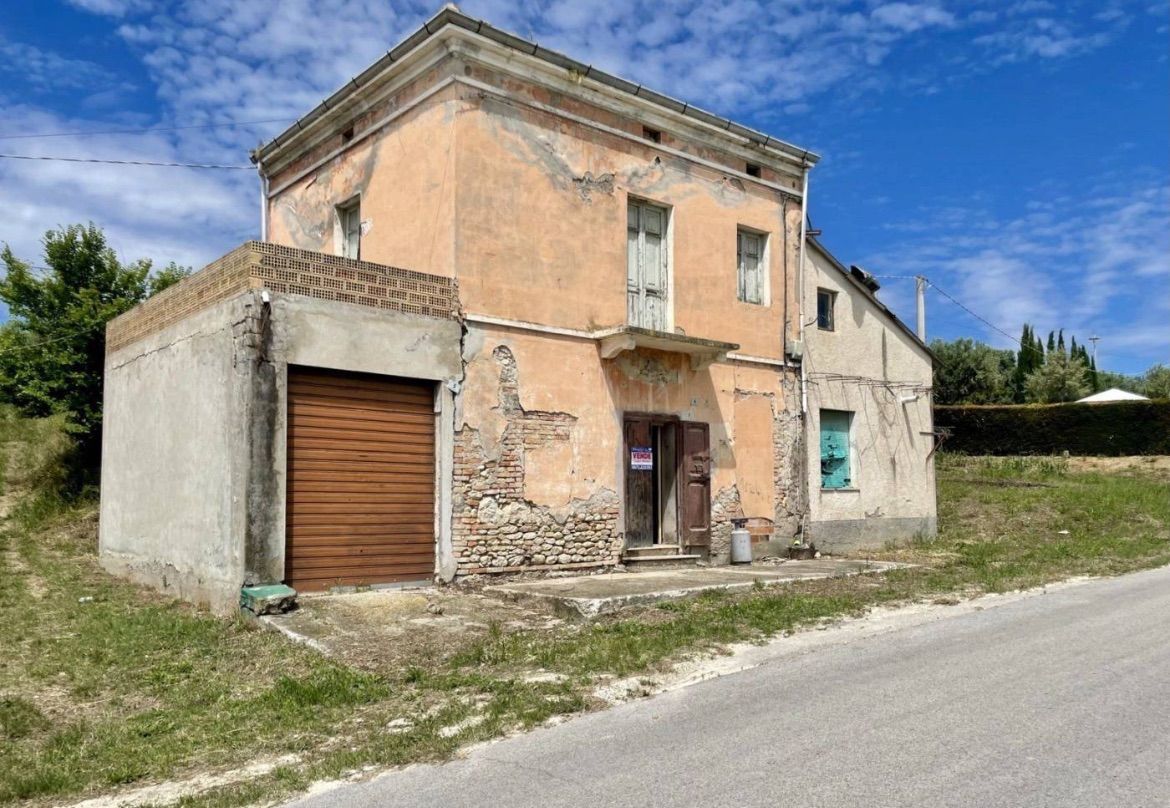 Two-storey rendered villa in Abruzzo with a concrete garage extension, cracked peach render, exposed masonry, shuttered upper windows and a narrow road in front.